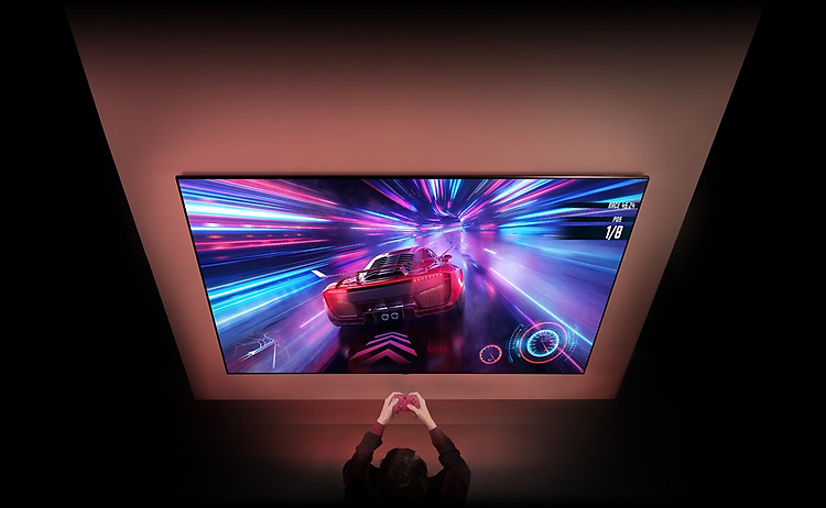 Có một chiếc TV lớn trên tường và bạn có thể nhìn thấy màn hình trò chơi đua xe trên màn hình. Trước TV, bạn có thể thấy bàn tay và bộ điều khiển của người tập trung vào trò chơi.