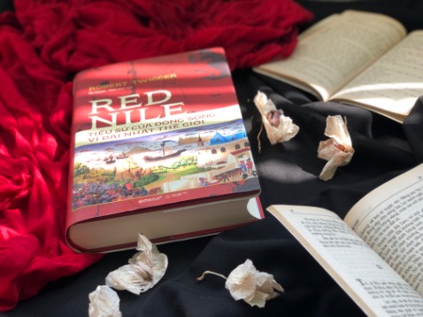 Nhà báo Trương Anh Ngọc viết trong lời giới thiệu:

“Sách rất dày, nhưng đọc vô cùng cuốn hút, không phải bởi tác giả đã viết rất duyên, mà còn vì con sông vĩ đại nhất thế giới ấy có biết bao câu chuyện để kể, và khi độc giả đã ôm lấy cuốn sách rồi, sẽ không muốn buông xuống nữa, vì dường như họ đang thấy lịch sử các vùng đất, các dân tộc, các sự kiện kéo dài mấy nghìn năm như một thước phim quay chậm trước mặt họ”. Đọc những lời mở đầu đó, tôi vừa tò mò và vô cùng hứng khởi để khám phá về Red Nile.
.

Lật giở từng trang sách, ta đắm chìm vào câu chuyện về con sông kì vĩ không chỉ về mặt tự nhiên mà còn cả những câu chuyện lịch sử đôi bờ nước chảy.

Trước tiên, hãy cùng Robert Twigger, lướt qua mặt nước sông, tìm về khởi nguồn sông Nile, qua hồ rộng, thác cao, qua đầm lầy Sudd rộng lớn rồi đến hồ No, đi thêm đoạn nữa ta sẽ bắt gặp nơi Nile trắng già cỗi gặp Nile xanh dữ dội và hợp thành Nile đỏ. Từ đây, con sông được hợp nhất. Uốn lượn qua những kim tự tháp lớn nhỏ của các Phraol đen, băng qua đập Aswan, qua đền Luxor diễm lệ rồi tiến vào thủ đô Cairo của Ai cập. Những toà nhà, những công trình mọc lên hai bờ sông, Nile tiếp tục vượt qua đập nước đầu tiên bắc ngang qua nó, Đập Delta, rồi dần dần tiến ra biển. 

Theo dòng nước, ta còn được lắng nghe những câu chuyện về thảm thực vật trù phú, về những kẻ cư dân nguy hiểm như khỉ đầu cho, hà mã, cá sấu và cả những vị thần dựa trên những con vật này. Ta được nghe kể những nỗ lực của con người trong việc chuyển hướng dòng sông, xây dựng đập nước để đảm bảo nước tưới tiêu quanh năm cho đôi bờ. Nhưng, thiên nhiên luôn khắc nghiệt và con người chẳng thể lường trước được mọi việc. Chính việc xây đập nước đã tạo môi trường thuận lợi cho muỗi, ốc sên… sinh sôi, truyền thêm bệnh dịch cho con người. Bên cạnh đó, sông Nile - con sông duy nhất chảy qua sa mạc Sahara vẫn như bao con sông khác. Khi đối mặt với cái nóng thiêu đốt, với gió mạnh cũng bị bốc hơi. Từ khi đập Aswan được xây dựng, những đợt lũ hàng năm không ồ ạt hung hãn tràn về xả bớt muối. Vậy là tấm áo ướt mồ hôi ấy không bao giờ được giặt và các dòng kênh phụ càng mặn hơn.

Cũng chính con sông ấy đã ban tặng cho vùng đất nơi nó chảy qua một món quà quý giá- giấy cói- nó đã giúp cho việc sử dụng chữ viết của con người trở nên riêng tư, hiệu quả và dễ vận chuyển hơn. Các mệnh lệnh và chỉ thị viết trên giấy cũng giúp con người cùng lúc dễ kiểm soát được nhiều hơn một thành phố. Thế rồi nhà nước đầu tiên do Ai Cập cổ đại định hình và giấy cói đóng một vai trò quan trọng. Cũng từ đó, lần theo dòng chảy của lịch sử, tác giả đã kể cho ta nghe những mẩu chuyện về các vị thần Ai Cập cổ xưa, về tình yêu và lòng thù địch, về kim tự tháp và những vị Pharon đầu tiên. Ta còn được nghe đến câu chuyện gay cấn về tai hoạ thắm đỏ, thích thú khám phá cuộc đời đầy thăng trầm và cũng lắm tai tiếng của vị Pharaon cuối cùng Cleopartra hay biết đến những người con kiệt xuất của Ai Cập cổ đại.

Màu đỏ của sông Nile chứa phù sa làm cho đất nước đôi bờ  trù phú và thịnh vượng, màu đỏ máu của những vị vua, những con người lưu lạc đến vùng đất này, màu đỏ của cả những tai hoạ đã góp phần làm nên thay đổi lịch sử của quốc gia nơi con sông uốn lượn chảy qua.

Có người đến và có những người đi, có câu chuyện của những kẻ xâm lấn và những công trình còn để lại, có những con đập minh chứng cho nỗ lực của con người trong việc điều tiết chuyển hướng dòng sông, có cả máu và nước mắt đã chảy trên Nile kì vĩ. Con sông chưa bao giờ ngừng biến đổi nhưng Nile vẫn luôn là nó, có lúc ồn ào và mạnh mẽ, đôi khi lặng lẽ chứng kiến bao đổi thay đến tận bây giờ.

Cuốn sách dài nhưng không hề khiến cho độc giả mỏi mệt bởi cách viết lôi cuốn, hấp dẫn về một chủ để mang tên tiểu sử.Robert Twigger đã làm cho một kẻ gà mờ về tự nhiên và lịch sử như mình vừa đọc vừa trầm trồ thán phục về câu chuyện của Nile.